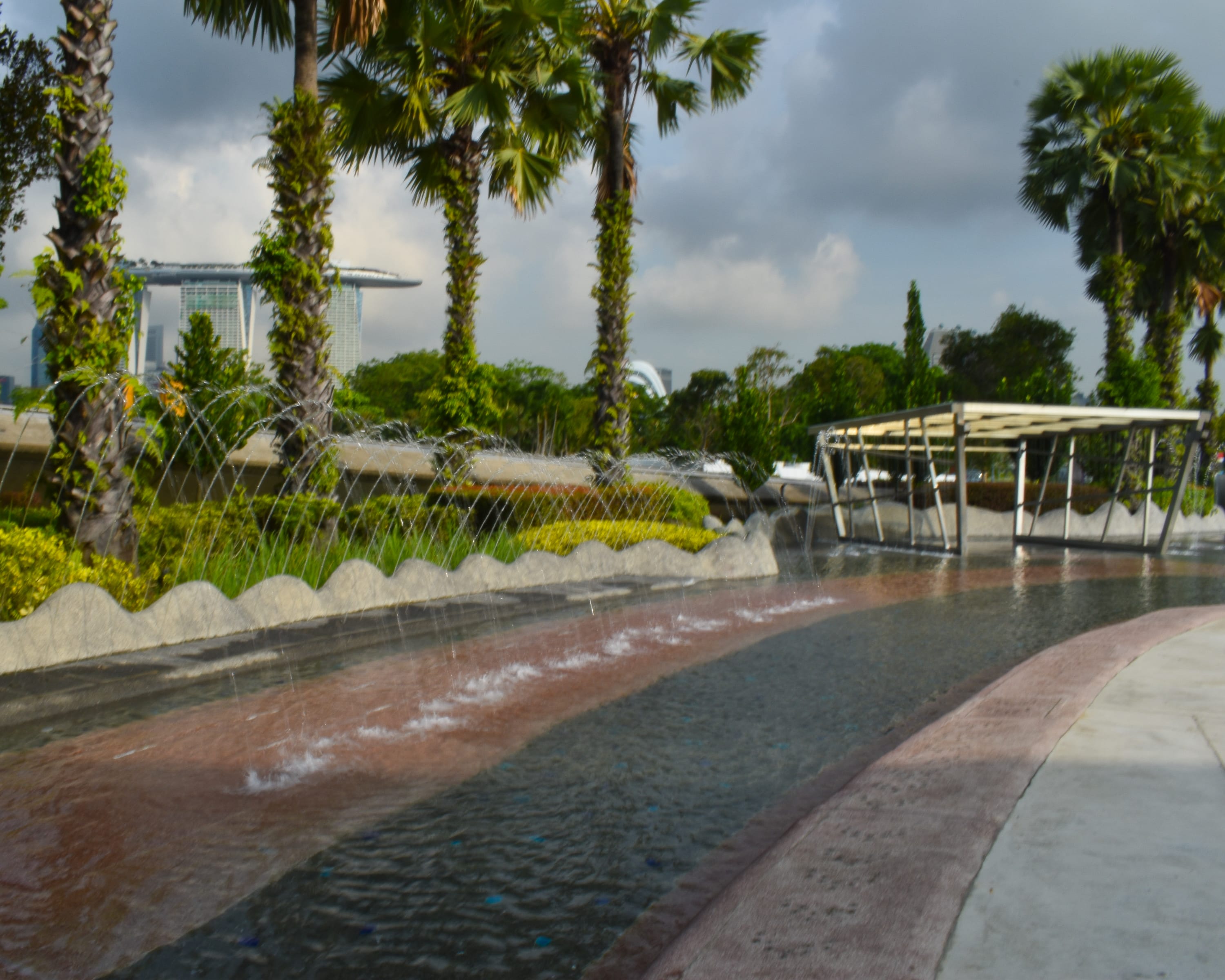 Curved water channel with arcing fountains, palm trees, and a covered walkway under cloudy sky.